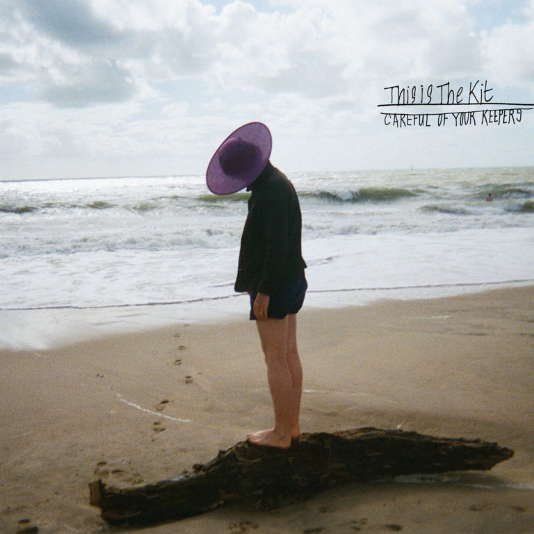 Cover des Albums „Careful of Your Keepers“ von This Is The Kit – Person mit lila Hut steht barfuß auf einem Treibholzstamm am Strand, Kopf gesenkt, Meer im Hintergrund.