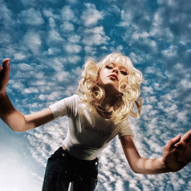 Maisie Peters blickt in die Kamera von unten, mit blond gelocktem Haar und „Father Figure“-Shirt vor dramatisch bewölktem Himmel – Cover von „The Good Witch“.