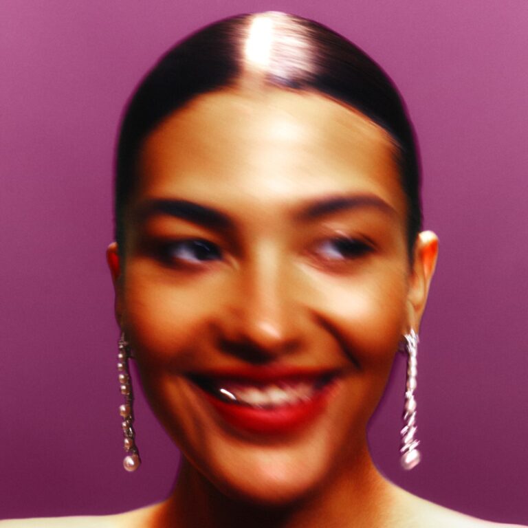 Porträt von Olivia Dean mit verschwommenem Lächeln vor violettem Hintergrund, glänzende Haare und elegante Perlenohrringe, Albumcover zu „Messy“.