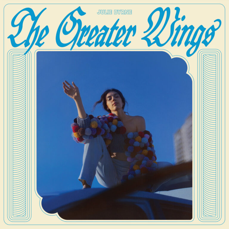Cover des Albums „The Greater Wings“ von Julie Byrne mit der Musikerin auf einem Auto sitzend vor tiefblauem Himmel, umrahmt von Retro-Typografie.