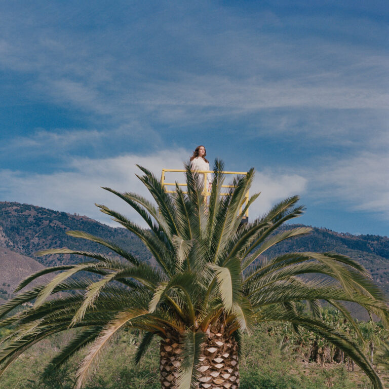 Jessy Lanza steht auf einer hohen Aussichtsplattform über einer Palme, vor strahlend blauem Himmel und Bergkulisse – Covermotiv zu Love Hallucination.