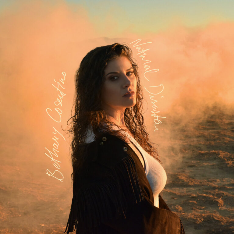 Bethany Cosentino steht in der Wüste vor orangem Dunst, trägt schwarzen Fransenmantel und blickt ernst in die Kamera – Albumcover zu Natural Disaster.