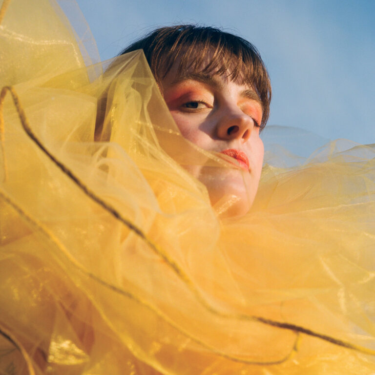 Nahaufnahme von Madeline Kenney in gelbem Tüll gehüllt, mit intensivem Blick und warmem Licht – Covermotiv zu A New Reality Mind.