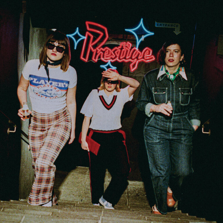 Albumcover von Girl Rays „Prestige“ zeigt drei Bandmitglieder in Retro-Outfits beim Verlassen eines Clubs mit Neon-Schriftzug „Prestige“ – stilvoll und nostalgisch.