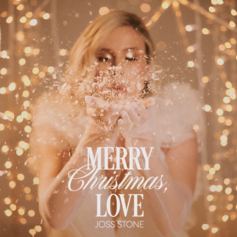 Joss Stone in weichem Licht vor goldenem Bokeh-Hintergrund, wie sie Schneeflocken in die Kamera pustet – Cover des festlichen Albums Merry Christmas, Love.
