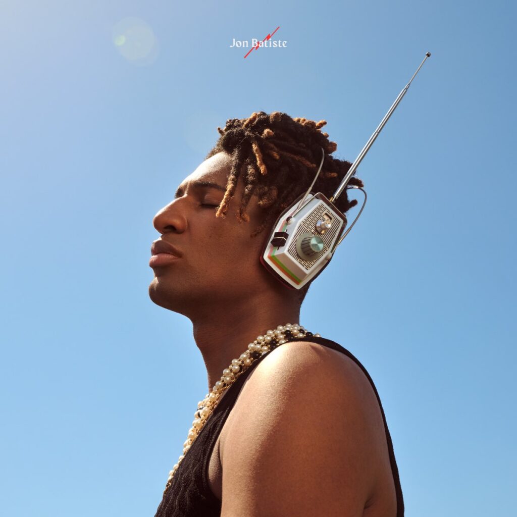 Jon Batiste mit geschlossenen Augen unter blauem Himmel, trägt ein Retro-Radio als Kopfhörer mit Antenne, Perlenkette und minimalistisches Top – Cover des Albums World Music Radio.
