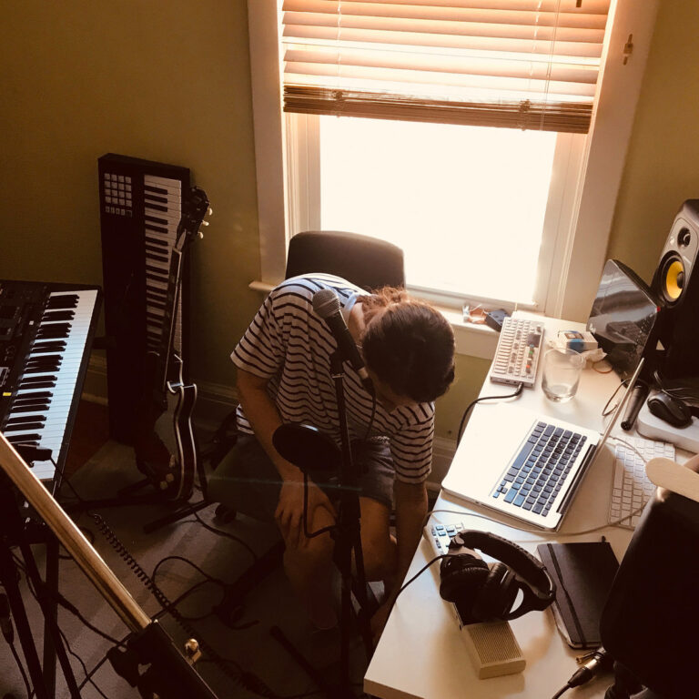Hannah Georgas sitzt in einem Heimstudio mit Mikrofon, Laptop, Kopfhörer und Keyboards vor einem sonnendurchfluteten Fenster, den Blick nach unten gerichtet – ein intimer Moment kreativer Selbstversunkenheit.