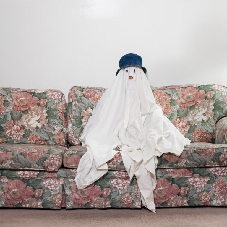 Ein mit einem weißen Laken bedecktes Wesen sitzt auf einem geblümten Sofa.