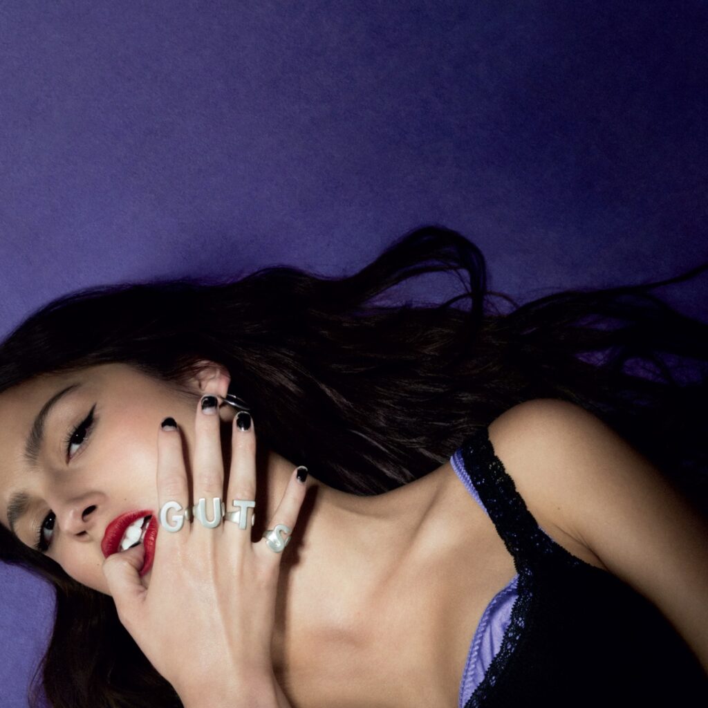 Porträt von Olivia Rodrigo mit „GUTS“-Ringen, liegend vor violettem Hintergrund, Teil des Albumcovers zu „GUTS“.