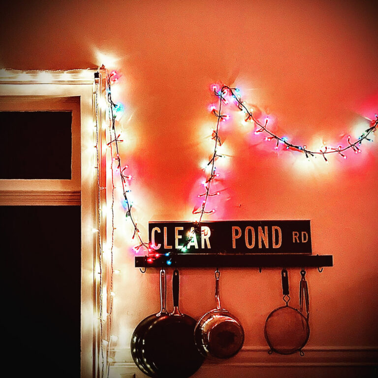 Albumcover von Kristin Hersh – Clear Pond Road: Eine Wand mit Lichterkette, Töpfen und einem nostalgisch beleuchteten Straßenschild in warmem Licht.