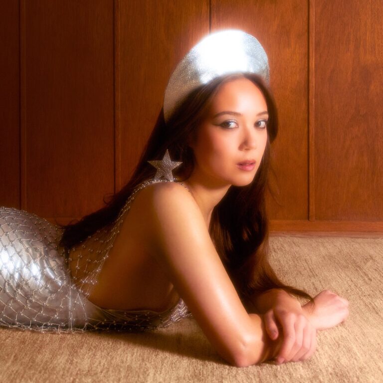 Laufey liegt in silbernem Kleid auf Teppich vor Holzvertäfelung, mit glitzerndem Kopfschmuck und Sternenohrring – Coverbild ihres Albums „Bewitched“.