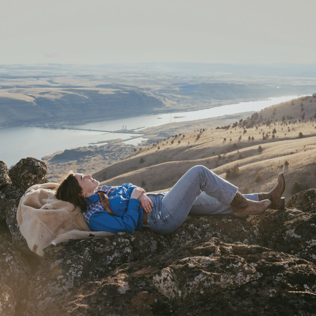 Margo Cilker liegt entspannt in Cowboystiefeln auf einem Felsen mit weitem Blick über ein Tal und einen Fluss – Sinnbild für das Album Valley of Hearts Delight.