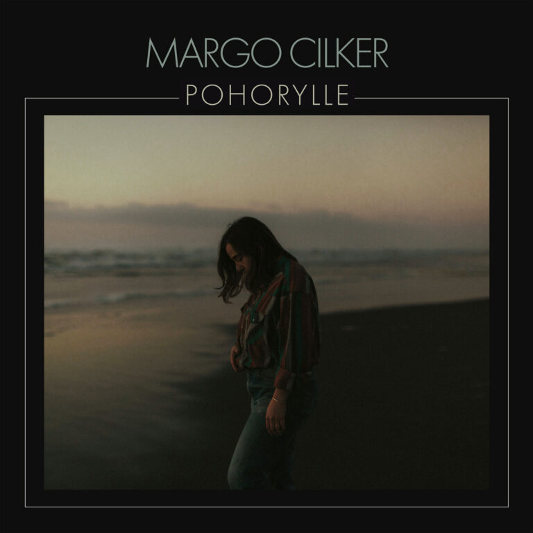Margo Cilker steht im Dämmerlicht am Strand, blickt nach unten, trägt kariertes Hemd – melancholisches Cover des Albums Pohorylle.