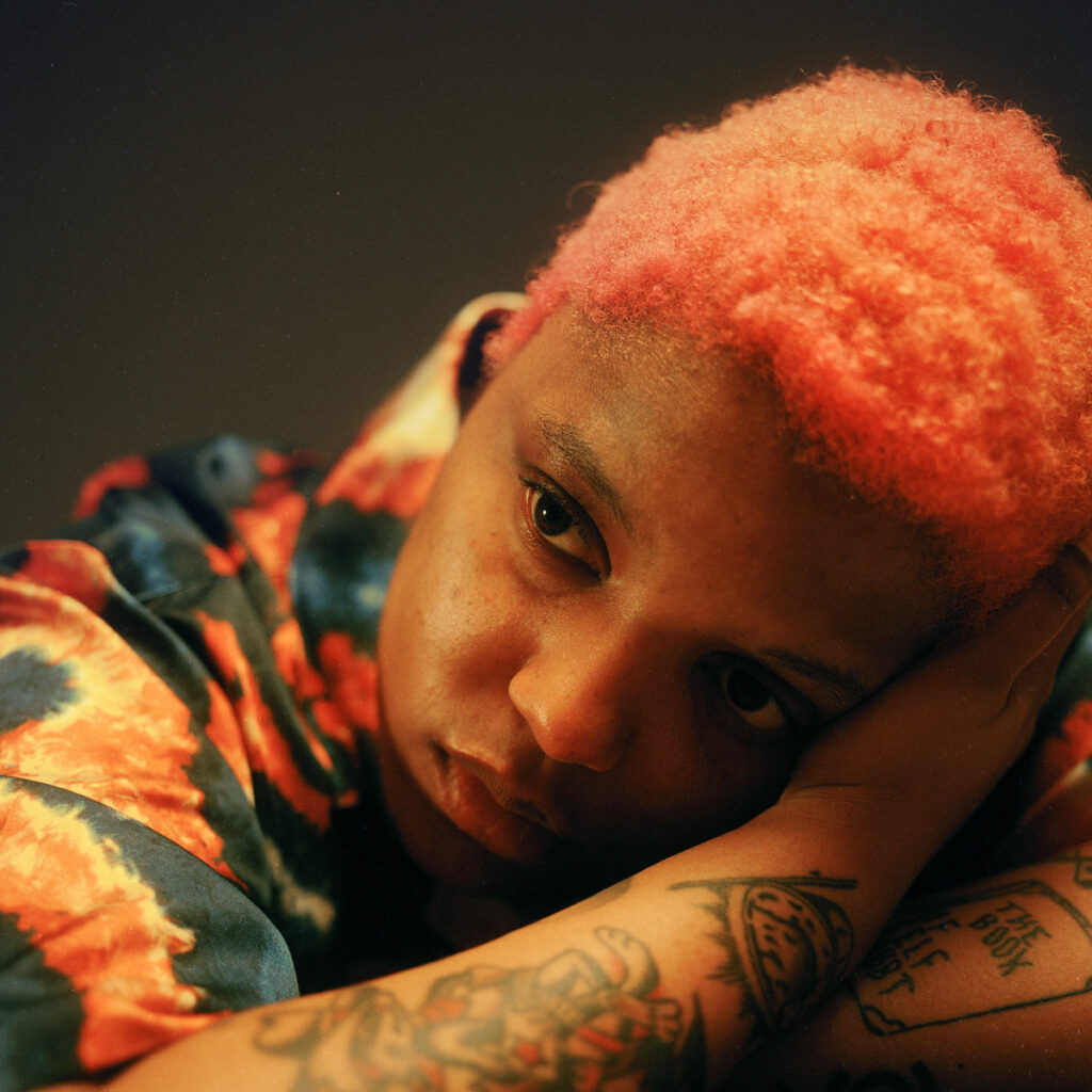 Porträt von Loraine James mit rosa gefärbtem Haar, tiefer Blick, Gesicht auf den Arm gestützt – ein Moment stiller Reflexion und emotionaler Nähe