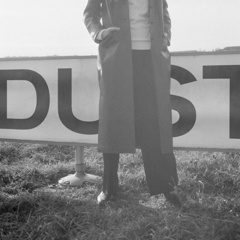 Person in langem Mantel steht vor einem Schild mit dem Wort DUST auf einer Wiese.