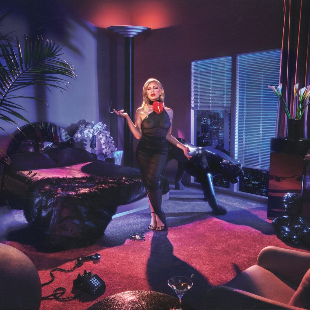 Slayyyter posiert in einem luxuriösen, neonbeleuchteten Schlafzimmer mit schwarzem Panther, Retro-Telefon und Cocktail – glamourös, provokant, selbstbewusst
