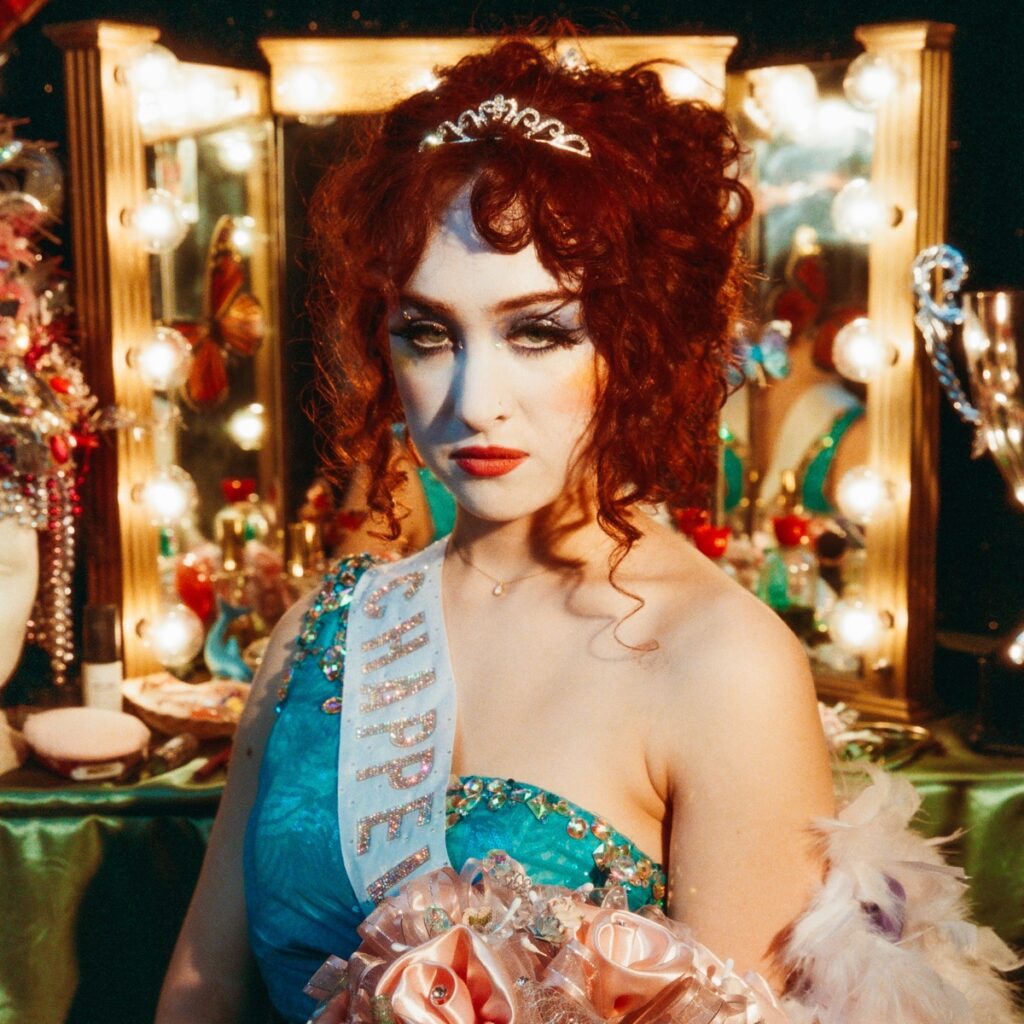Albumcover zu „The Rise and Fall of a Midwest Princess“ von Chappell Roan – sie posiert als traurige Schönheitskönigin vor beleuchtetem Schminktisch, mit Tiara, Schminke und Satin-Schärpe.