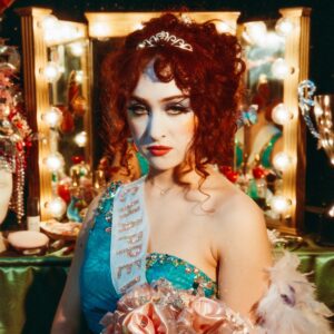 Albumcover zu „The Rise and Fall of a Midwest Princess“ von Chappell Roan – sie posiert als traurige Schönheitskönigin vor beleuchtetem Schminktisch, mit Tiara, Schminke und Satin-Schärpe.