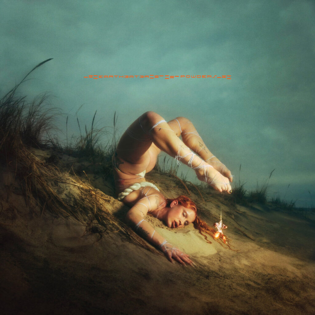 Albumcover zu „Powders“ von Eartheater – sie liegt in surrealer Pose am Sandhang, mit Körperkunst und Bindfäden umwoben, während an ihrem Haar eine kleine Flamme lodert.