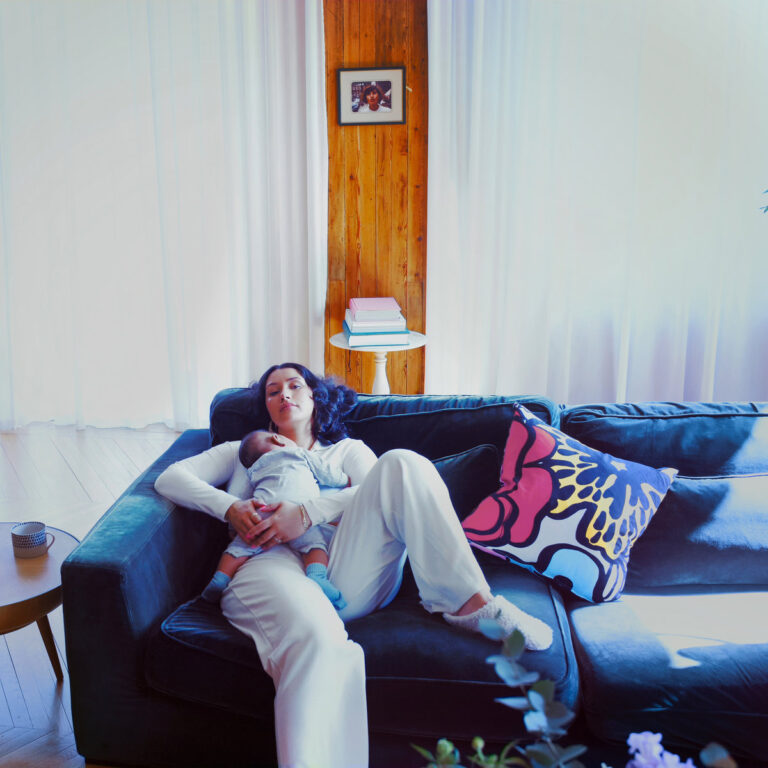 Cleo Sol liegt im weißen Pyjama mit Baby im Arm auf einem tiefblauen Sofa – ein intimer Moment zwischen Mutterschaft, Wärme und Erinnerung.