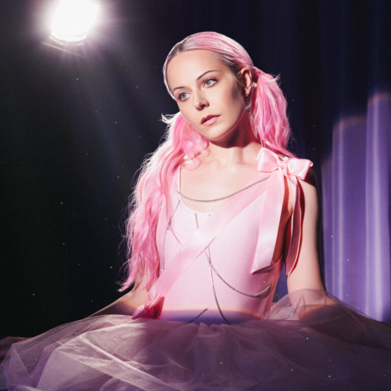 Pressebild zu Hannah Diamond – in rosa Tüll und Schleifen unter Spotlight, mit pinken Haaren und verträumtem Blick ins Leere, auf einer Bühne inszeniert.