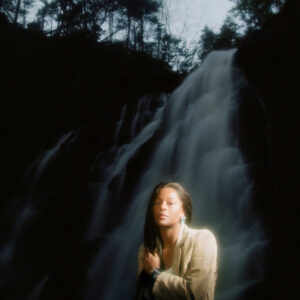 Porträt der Musikerin L'Rain vor einem mystisch beleuchteten Wasserfall im Dunkeln, mit verträumter Lichtstimmung und Naturkulisse.