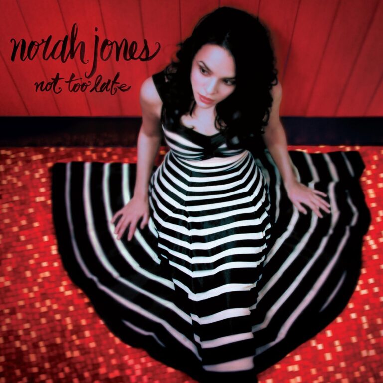 Norah Jones sitzt in einem rot beleuchteten Raum und trägt ein schwarz-weiß gestreiftes Kleid.