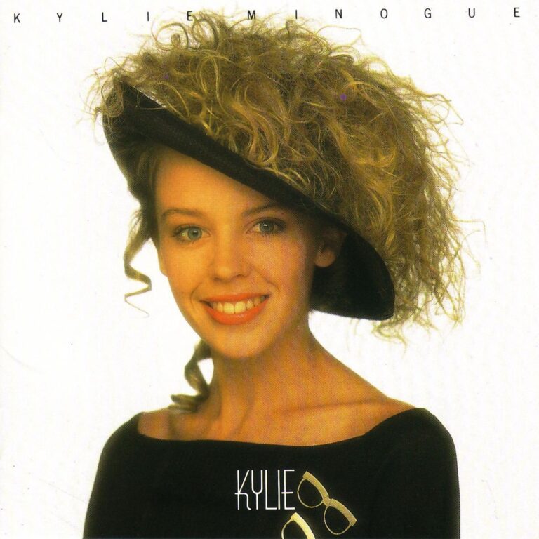 Kylie Minogue lächelt in einem schwarzen Oberteil mit Hut und Brille auf einem Albumcover.