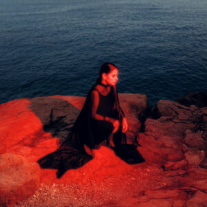 Albumcover von „Madres“ zeigt Sofia Kourtesis in schwarzem Outfit auf rötlich beleuchtetem Felsen vor dunklem Meer – atmosphärisch und introspektiv.