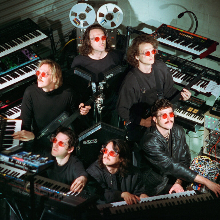 Pressebild zu „The Silver Cord“: King Gizzard & The Lizard Wizard in schwarzer Kleidung mit roten Brillen, umgeben von Vintage-Synthesizern und Kabeln.