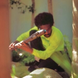 André 3000 sitzt mit Flöte im Freien, trägt ein neongelbes Shirt – stimmungsvolles, unscharf fotografiertes Coverbild seines Ambient-Albums „New Blue Sun“.