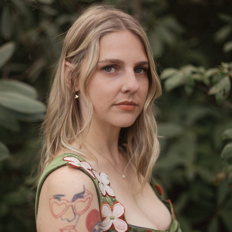 Porträt der Musikerin Dori Freeman vor grünem Blätterhintergrund, mit floraler Kleidung und sichtbarem Tattoo auf dem Oberarm.