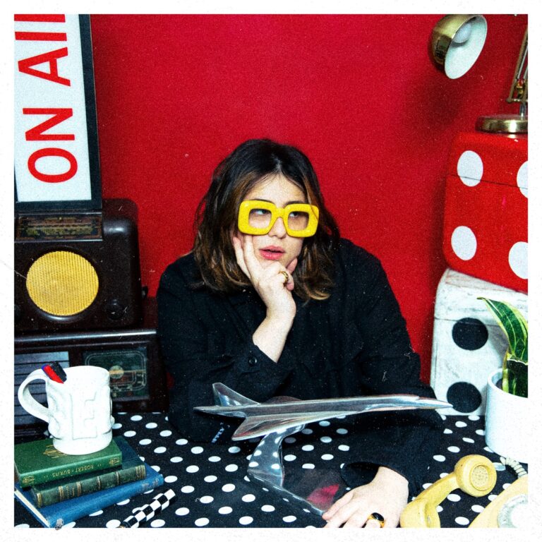 Belot sitzt mit übergroßer gelber Brille an einem Retro-Schreibtisch vor rotem Hintergrund – Cover ihrer EP „The Good Life“.
