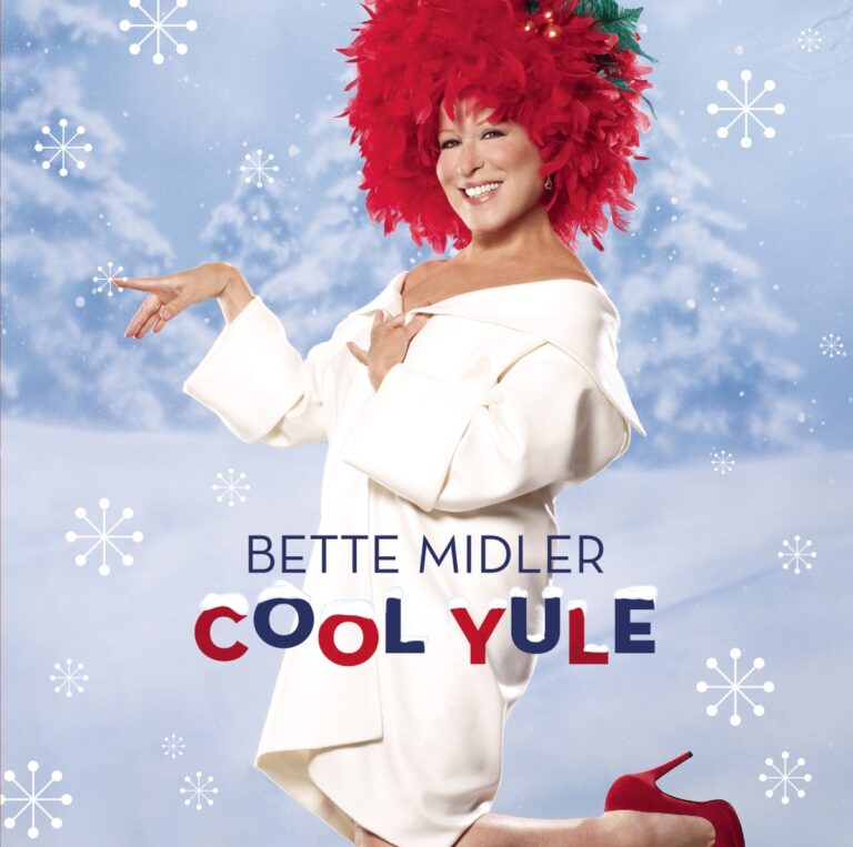 Bette Midler posiert in einem weißen Outfit mit rotem Haarschmuck vor einer winterlichen Kulisse.
