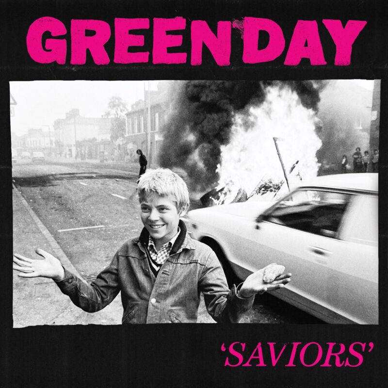 Albumcover von „Saviors“ von Green Day: Ein Junge steht lächelnd vor einem brennenden Auto in einer schwarz-weißen Straßenszene; darüber in grellem Pink der Bandname – ein ikonisches Sinnbild für anarchischen Spott und Pop-Punk-Attitüde.