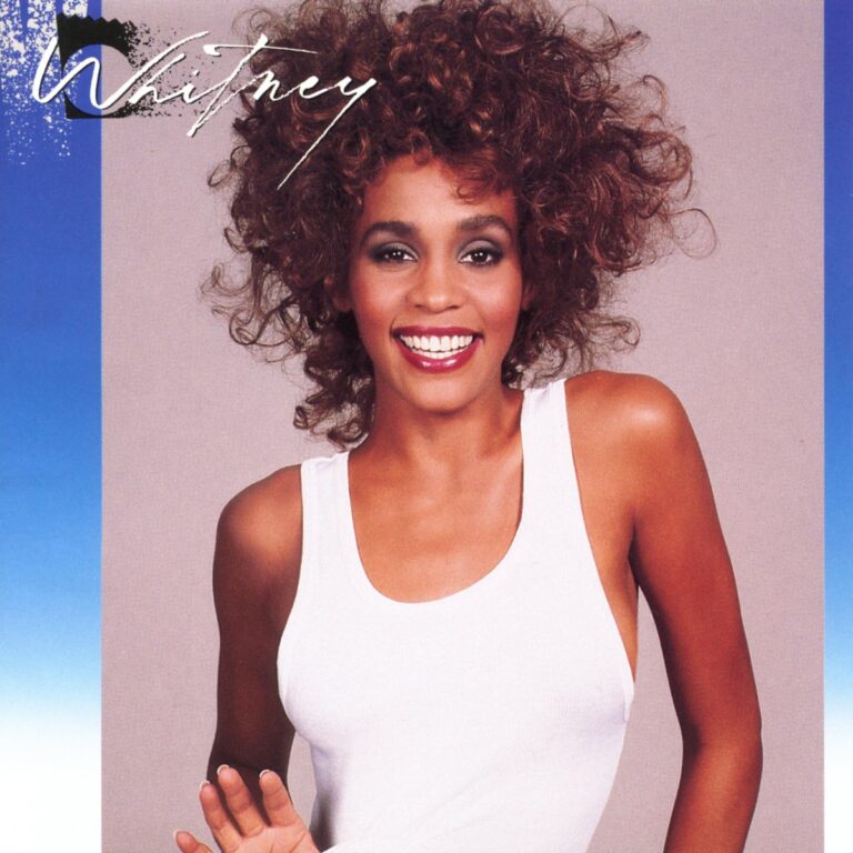 Whitney Houston lächelt in einem weißen Top vor einem blauen Hintergrund.