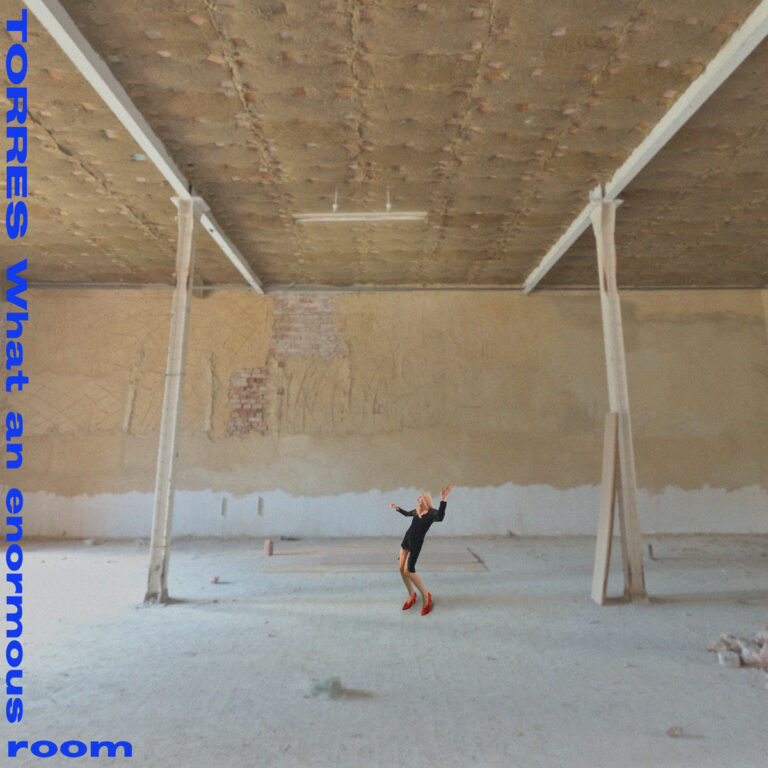Cover des Albums „What an Enormous Room“ von TORRES: eine Miniaturfigur in schwarzem Kleid mit roten Schuhen steht verloren in einem leeren, rohen Industrie-Raum – Sinnbild für emotionale Weite und intime Selbstreflexion.