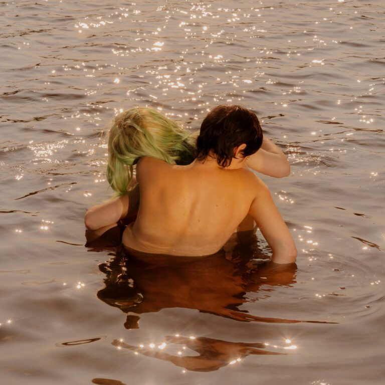 Zwei Oberkörper tauchen eng umschlungen in golden schimmerndem Wasser – ein sinnlich-intimer Moment, der das ambivalente Gefühlsspektrum von Katy Kirbys Album „Blue Raspberry“ visuell aufgreift.