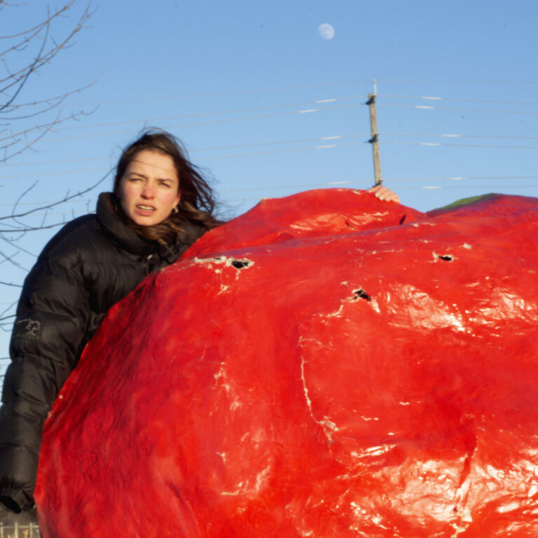 Albumcover von PACKS – Crispy Crunchy Nothing: Madeline Link lehnt im Daunenmantel an einer riesigen roten Pappmaché-Frucht, blauer Himmel, Mond, Strommast im Hintergrund.