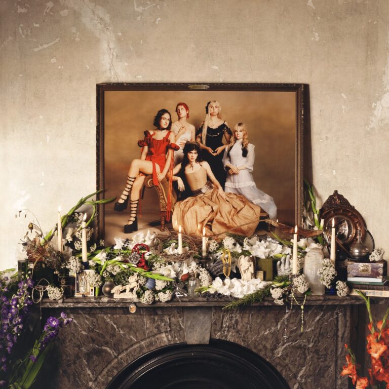 Cover von The Last Dinner Party – Prelude to Ecstasy: ein barockes Gruppenporträt auf einem Kamin, umgeben von Blumen, Kerzen und viktorianischen Dekorationen.