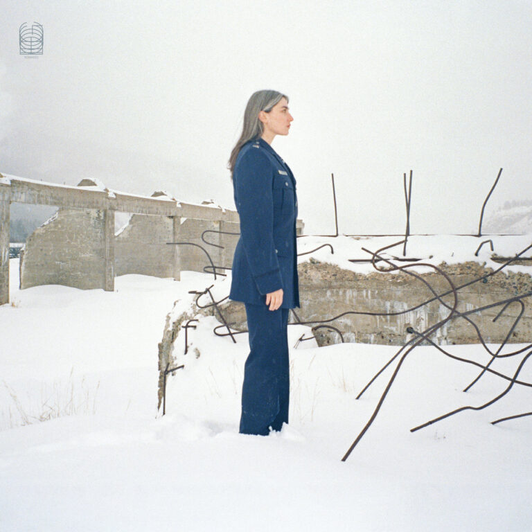 Cover von Kali Malone – All Life Long: die Künstlerin steht seitlich im Schneefeld vor einer verfallenen Ruine, gekleidet in eine blaue Uniform, umgeben von Beton und Stahl.