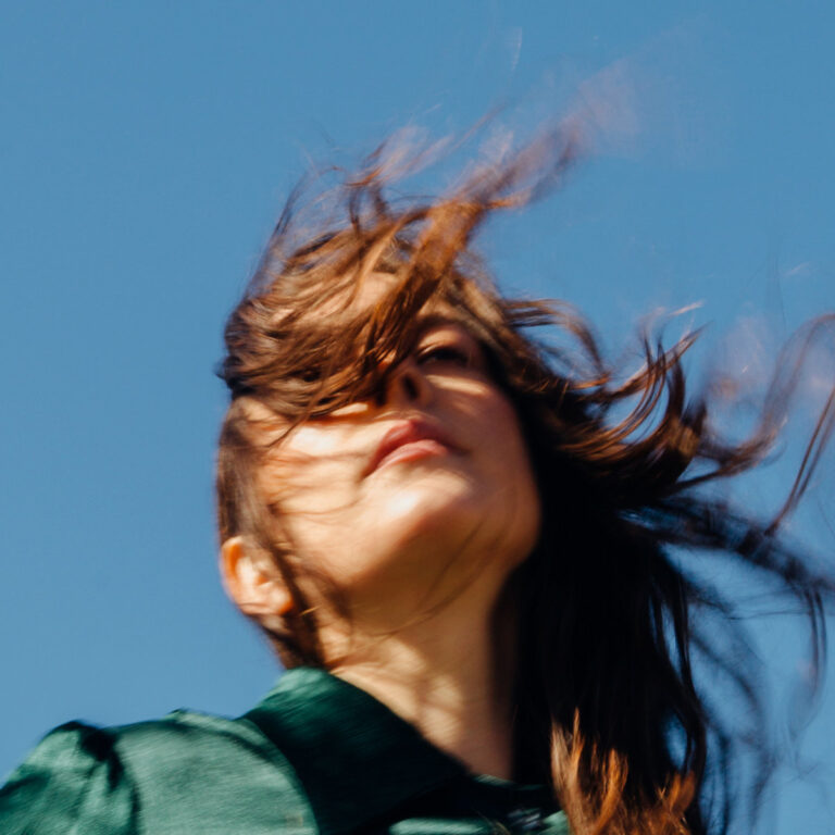 Madi Diaz mit vom Wind verwehtem Haar vor klarem Himmel – Albumcover zu „Weird Faith“, das ihre verletzliche, aber kraftvolle Musik visuell einfängt.