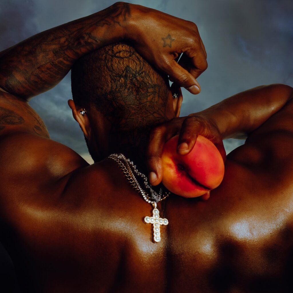 Cover von Usher – Coming Home: der tätowierte Rücken des Sängers mit Kreuzkette, erhobener Arm und Pfirsich in der Hand vor dramatischem Himmel.