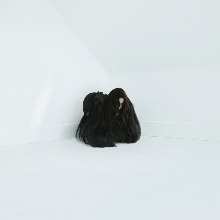 Person mit langen Haaren kauert in einer weißen Ecke.