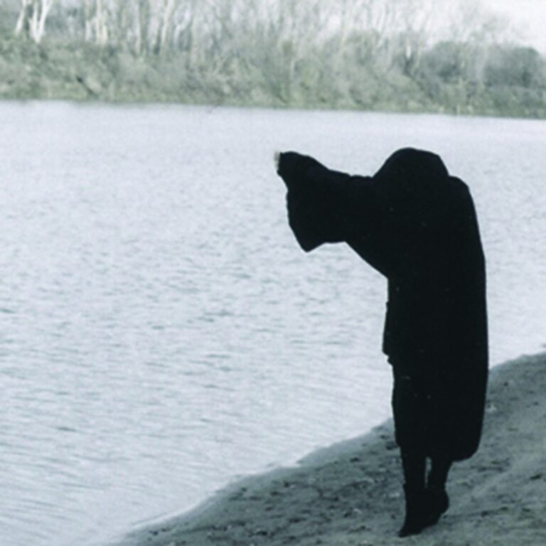 Person in schwarzem Umhang am Ufer eines Sees mit Bäumen im Hintergrund.