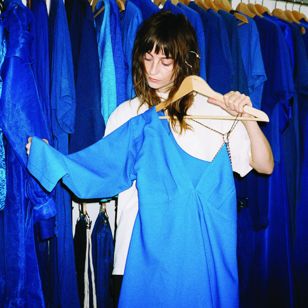Cover von Faye Webster „Underdressed at the Symphony“: Die Musikerin hält ein leuchtend blaues Kleid inmitten einer monochrom blauen Garderobe.