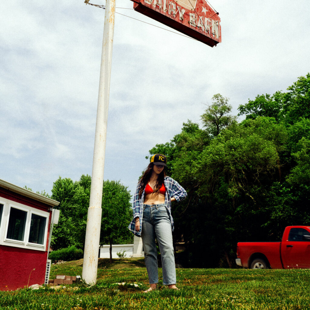 Albumcover von Waxahatchee – Tigers Blood: Katie Crutchfield barfuß unter einem verblassten Schild, in ländlicher Kulisse mit rotem Pickup und KC-Cap.