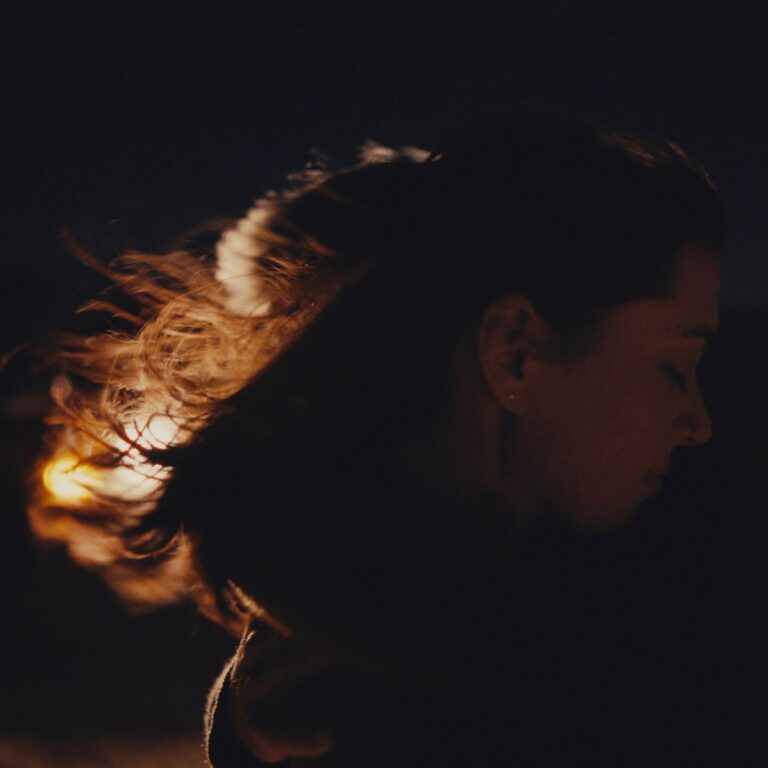 Seitenportrait von Lizzy McAlpine im Dunkeln, ihr Haar weht im warmen Licht, während sie nachdenklich den Blick senkt.