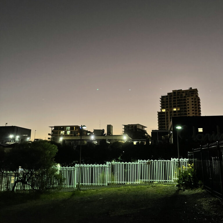 Dämmerungsaufnahme urbaner Skyline mit hell beleuchtetem Zaun im Vordergrund – Symbolbild zum Album „Final Summer“ von Cloud Nothings.