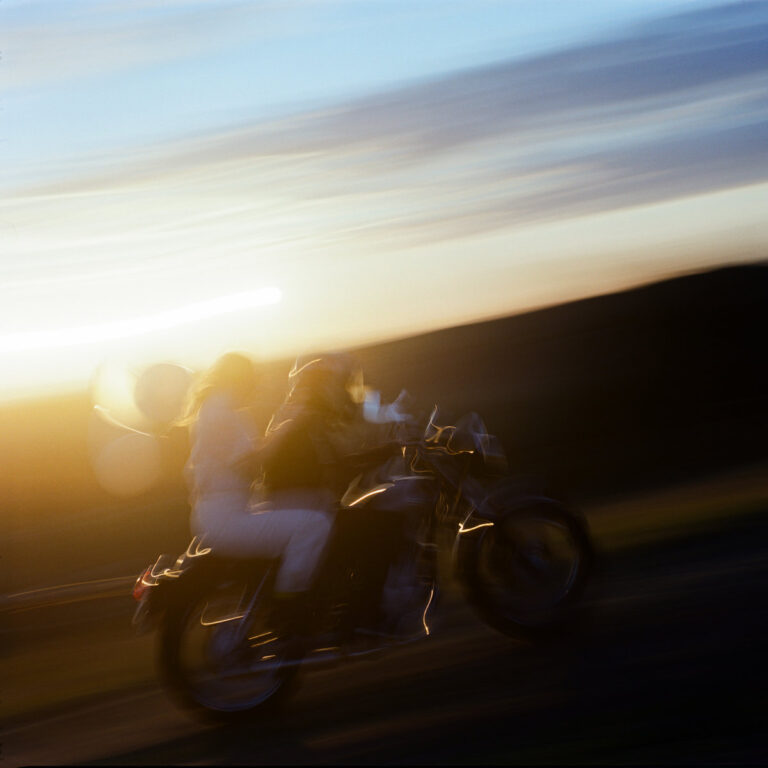 Verschwommene Aufnahme zweier Personen auf einem Motorrad bei Sonnenuntergang – ein Bild voller Tempo, Freiheit und emotionaler Aufladung.